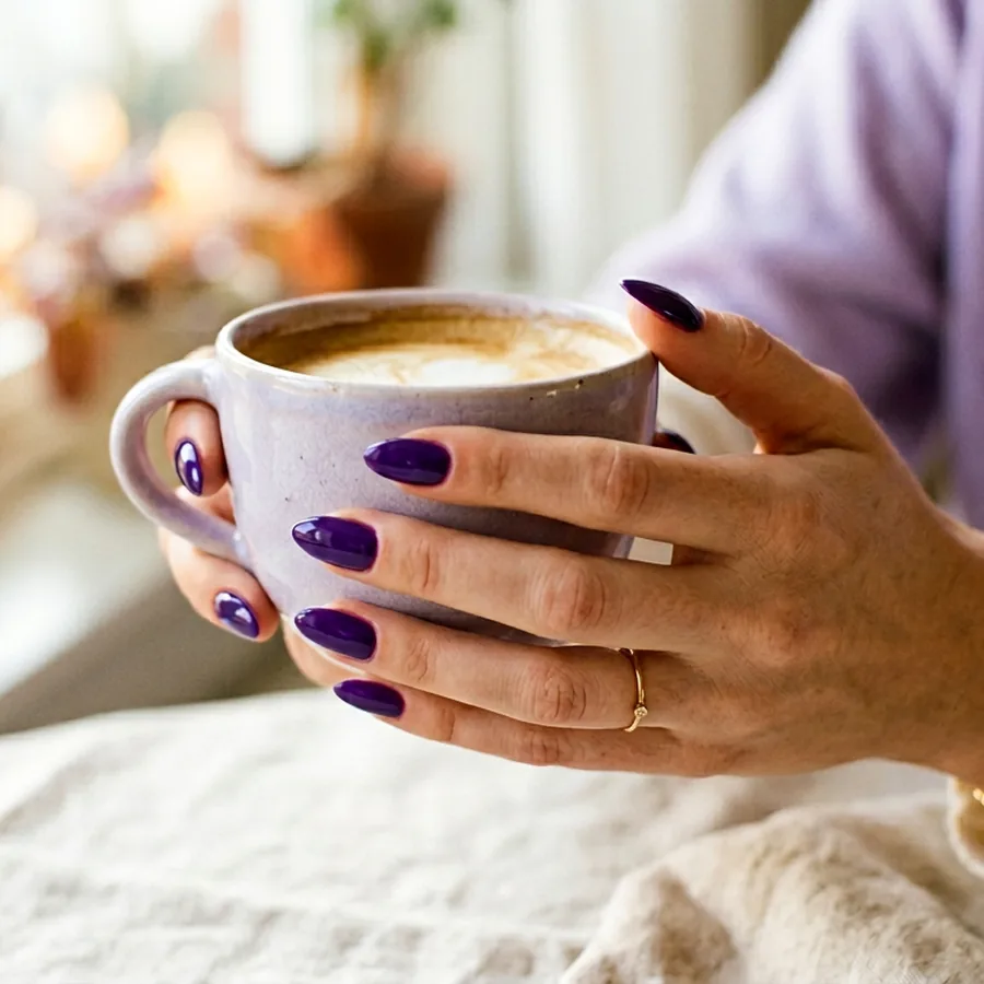 Glossy deep violet gel manicure on a hand holding a pale lilac ceramic coffee cup